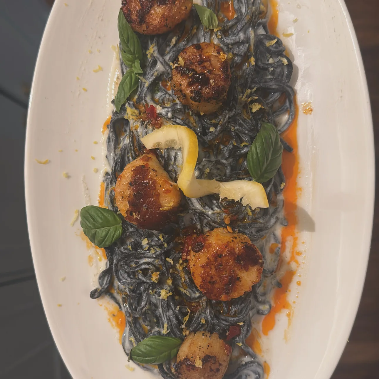 Seared scallops on squid-ink pasta with basil and a lemon twist, on a white oval platter