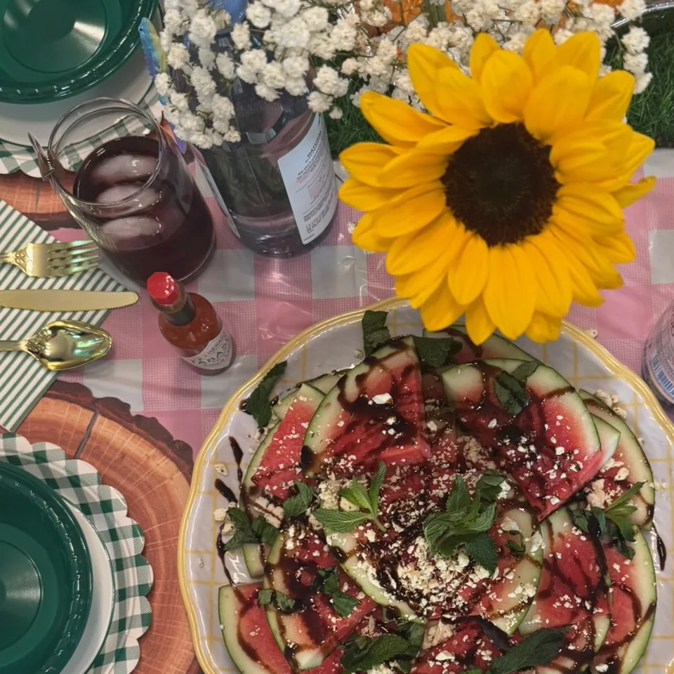 A watermelon and feta salad on a laid table — gingham cloth, green plates, gold cutlery and a sunflower